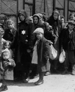 Black and white photo of Jews with Star of David patches sewn on their jackets stand in line near railroad cars during the Holocaust